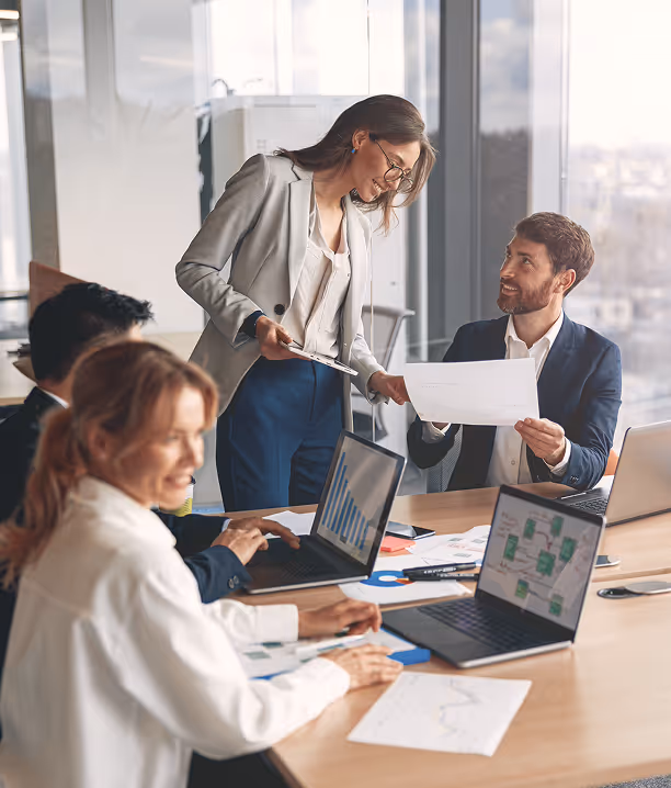 Four professionals collaborating in a modern office, discussing documents and working on laptops displaying charts and graphs.